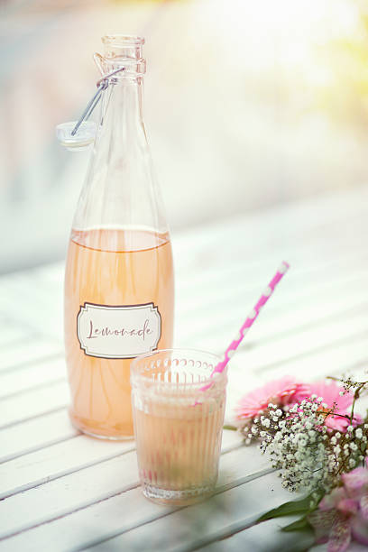 A bottle of pink lemonade with a matching glass and striped straw on a summer table.