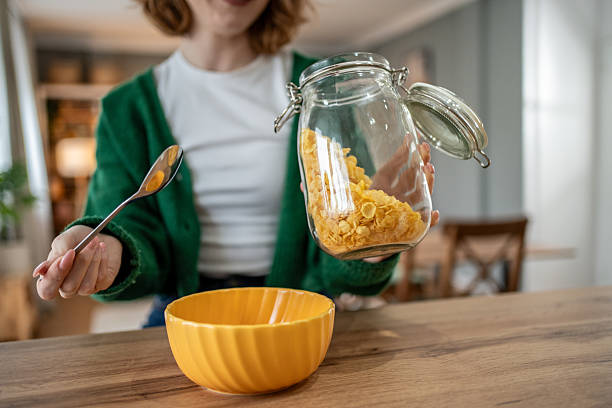 Pouring cornflakes from a glass jar into a vibrant yellow bowl, a woman prepares for a nutritious and enjoyable breakfast, embracing a healthy lifestyle in her cozy kitchen