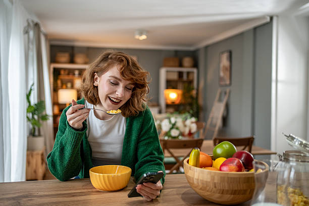 istock Happy young woman eating cereal and using smartphone in kitchen 2209612052