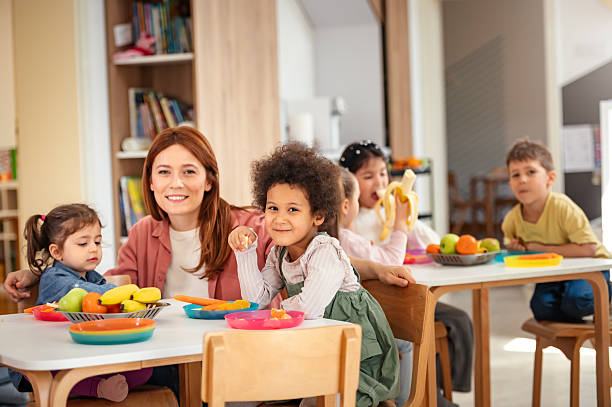 istock A diverse group of preschool children sits around a table eating healthy snacks while a teacher engages with them, promoting nutrition, inclusion, and social interaction in early education. 2209617480