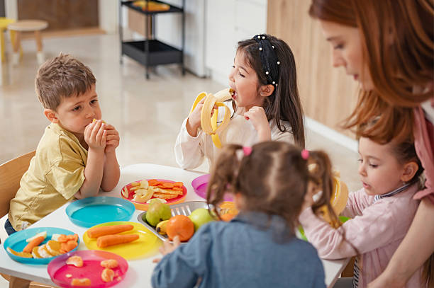 istock A diverse group of preschool children sits around a table eating healthy snacks while a teacher engages with them, promoting nutrition, inclusion, and social interaction in early education. 2209617556
