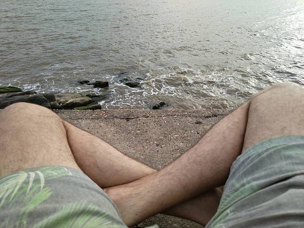 Rear view first person perspective man sitting by water with legs visible and  shorts.