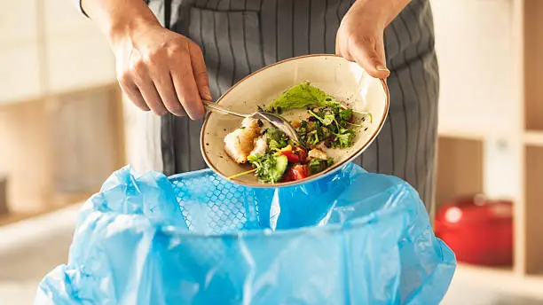 Woman scraping leftover food from plate into trash bin in kitchen Woman scraping leftover food from plate into trash bin in kitchen