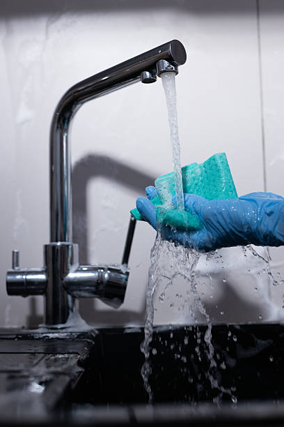 The image captures a hand wearing a blue glove holding a sponge under a gleaming kitchen faucet, demonstrating the process of cleaning in a contemporary kitchen environment.