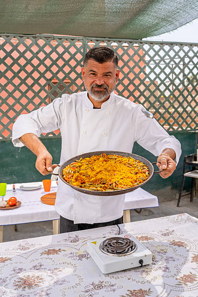 Chef proudly presenting a vibrant paella, freshly prepared outdoors on a sunny rooftop, showcasing the essence of spanish cuisine