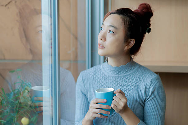istock Thoughtful Asian woman holding a blue coffee mug while looking out the window, reflected in glass, enjoying a peaceful indoor moment in natural soft light 2209656240