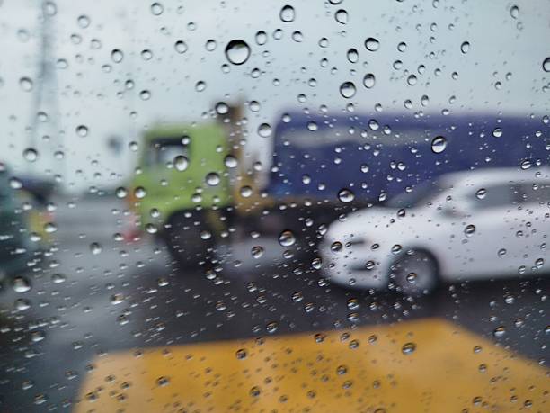 Raindrops cling to the window, blurring the view of vehicles stuck at a road junction. A green truck and a white car appear hazy behind the curtain of rain, creating a moody yet artistic scene.