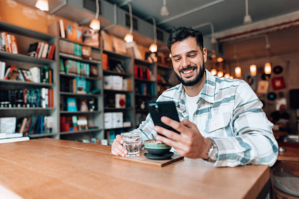 istock Young man smiling and using mobile phone in a bookshop cafe 2209712088
