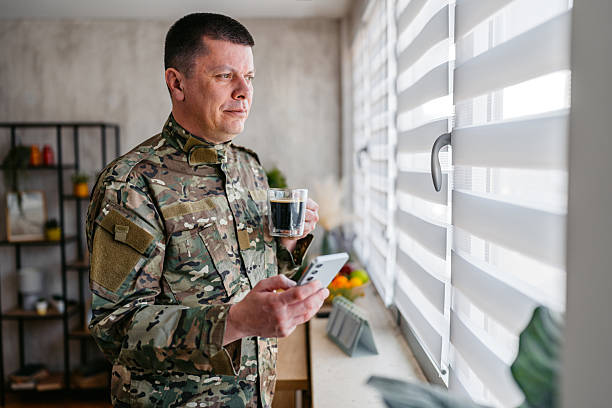 Pensive mid-adult soldier in a military uniform drinking coffee and using phone while looking out the window in the living room.