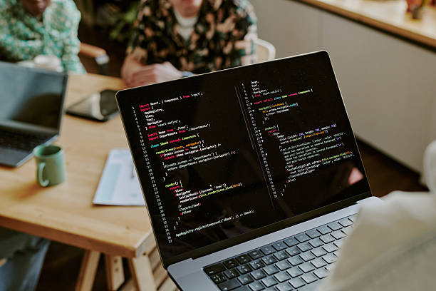 Over shoulder view of silver laptop with black screen full of code lines in hands of unrecognizable speaker giving presentation in front of team