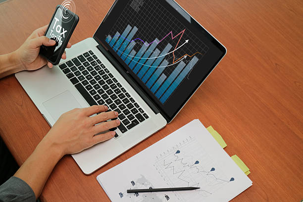 Hands working on a laptop and holding a smartphone with charts and graphs displaying tax and tariffs growth trends.
