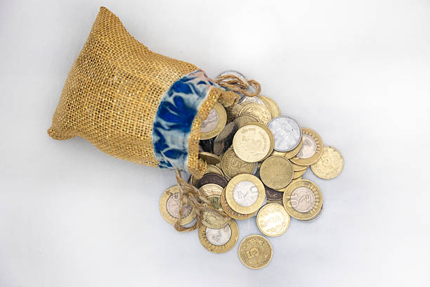 A burlap sack with a patterned top spills coins of various denominations over a white background, symbolizing wealth, savings, and financial accumulation.
