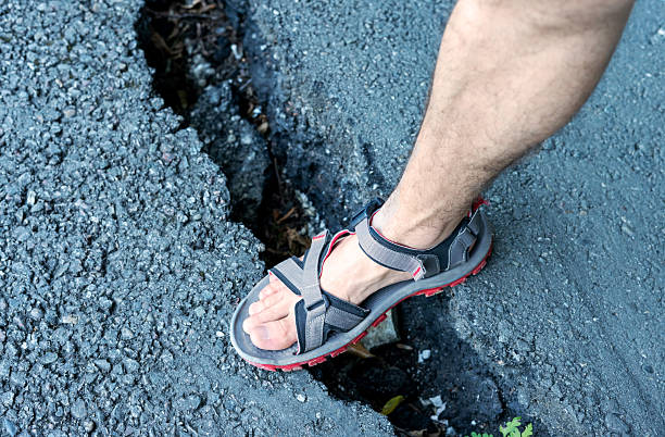 istock Foot in grey sandal steps into crack on an asphalt road, infrastructure damage, posing potential hazards for pedestrians Outdoor setting with visible gravel 2209734707