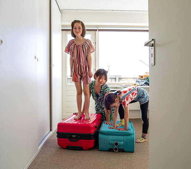 Real moments of family packing their suitcases and bags in their homes to prepare for an upcoming trip. Children trying to close the suitcase, mother helping in the background.