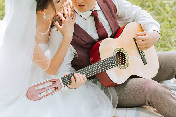 A couple is sitting on a blanket and playing a guitar. The woman is wearing a white dress and the man is wearing a red vest