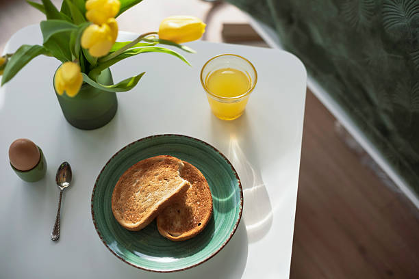 Cozy breakfast scene with a fresh juice, toast, and a soft-boiled egg on a white table, next to a vase of yellow tulips in a bright, minimalist home interior. Natural morning light and warm tones create a peaceful and inviting atmosphere.