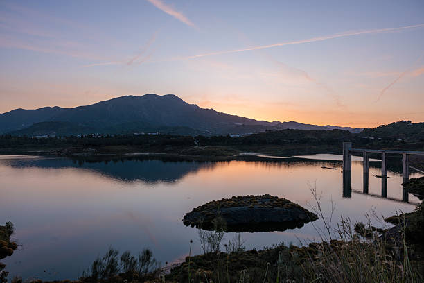 Tranquil lake mirroring mountains and colorful sunset sky with concrete bridge structure, creating a peaceful and scenic landscape