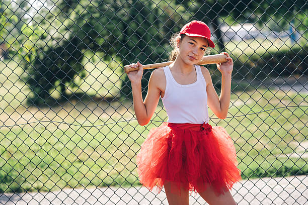 A creatively dressed girl in a red tutu and cap poses with a baseball bat, showcasing her playful spirit against a lively background that highlights her vibrant energy.