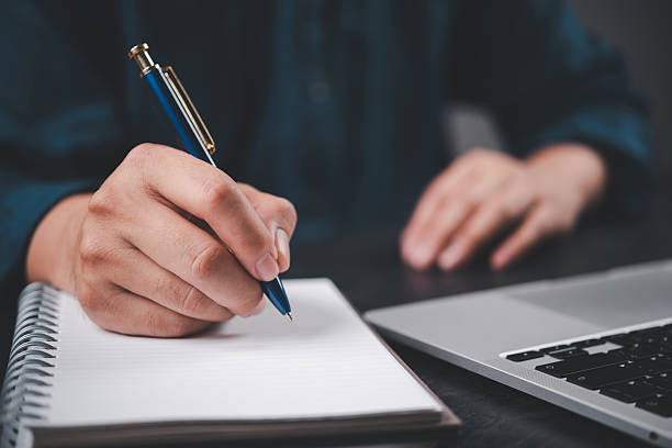 istock Close-up hand writing on notebook with pen next to laptop. Education, business planning, journaling setup with modern workspace productivity and creative thinking. 2209760281