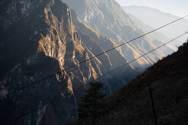 This photo, taken in the late afternoon in Tiger Leaping Gorge, Yunnan, China, showcases sunlight bathing steep mountain ridges, highlighting their contours and geological textures. The layered peaks recede into the distance under a soft atmospheric haze, while dark power lines cut diagonally across the frame—an unmistakable sign of human infrastructure. The image blends sublime natural beauty with subtle signs of development, evoking the quiet tension between modern access and remote wilderness in this dramatic part of southwestern China.