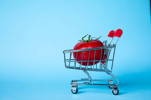 A vibrant red tomato sits in a small shopping cart against a blue background, symbolizing grocery shopping and healthy eating concepts in a playful and imaginative way.