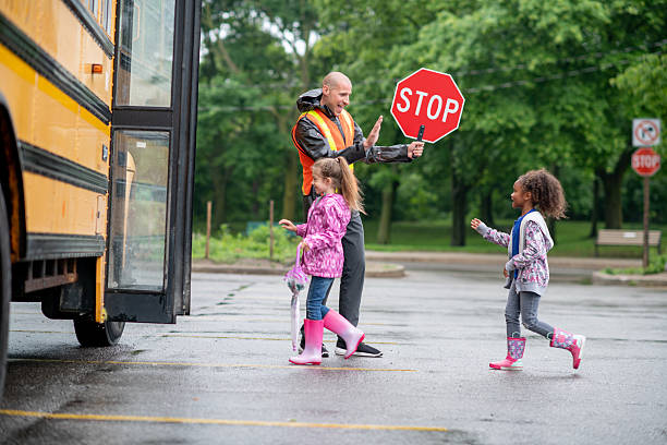 A school crossing guard directs two young children, ensuring they cross safely to board a school bus on a rainy day.