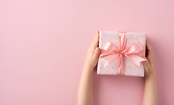 istock Delicate pink gift box tied with a satin ribbon held by hands, on plain backdrop 2209795541