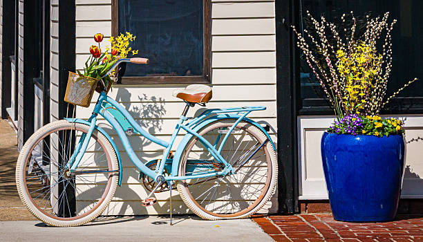 The basket of a bicycle is filled with springtime tulips and forsythia branches as it is parked on a sidewalk next to a large blue urn with pussy willows, forsythia and pansies