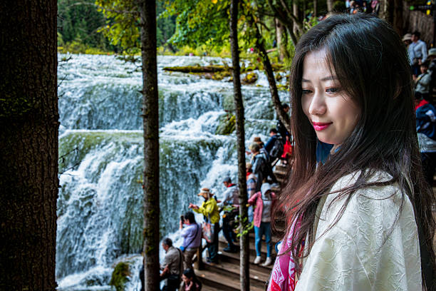A woman stands in front of a waterfall, dressed in a white outfit with a red scarf, showing a bright smile against the stunning natural backdrop