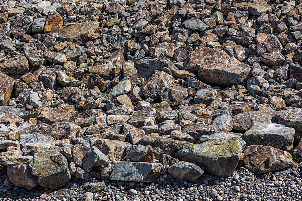 Heap of natural, irregularly shaped stones and rock fragments of different sizes scattered on a surface, illuminated by strong daylight creating shadows and highlighting textures and earthy tones of brown, gray, and hints of green.