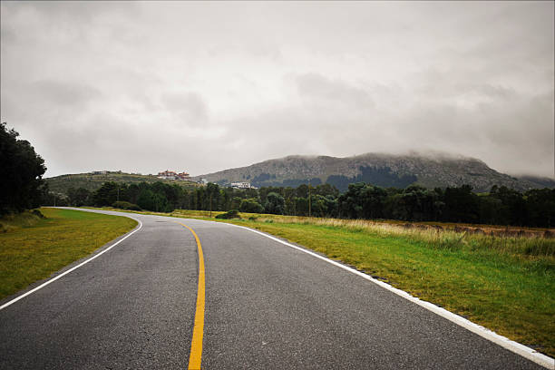 Under a heavy gray sky, a quiet road curves gently into the distance, leading toward misty hills cloaked in low clouds. The moody atmosphere, softened by the muted greens of the grass and trees, invites contemplation. On the horizon, a cluster of buildings nestles into the landscape, dwarfed by the looming, fog-shrouded mountains behind them. It’s a scene that whispers of slow journeys, hidden destinations, and the peaceful solitude of a road less traveled.