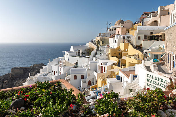 Santorini, Greece. April 15th, 2023: A beautiful view of the iconic whitewashed buildings of Santorini, with the Aegean Sea stretching out to the horizon. The vibrant flowers in the foreground add a pop of color to the scene, while the clear blue sky and calm sea enhance the peaceful atmosphere of the island.