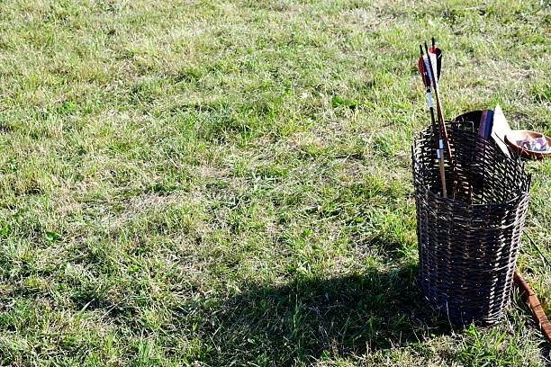 A close up on a set of arrows with decorative feathers on their backs inserted into a wicker basket, laying next to a lush field, meadow, or pastureland seen in Poland in summer