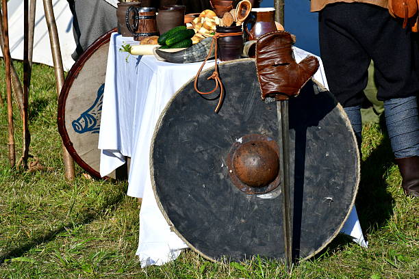 A close up on a set of medieval or Scandinavian shields located next to a glove, a sharp sword and a table full of medieval meals and drinks seen in summer during a historic fair in Poland