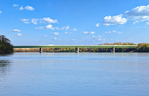The M0 motorway bridge forms a vital transportation link across the Danube River in the southern outskirts of Budapest, Hungary. The modern structure cuts a clean line above the calm blue waters, while the bright sky and scattered clouds create a serene backdrop for this essential piece of Hungarian infrastructure that connects different parts of the capital city.