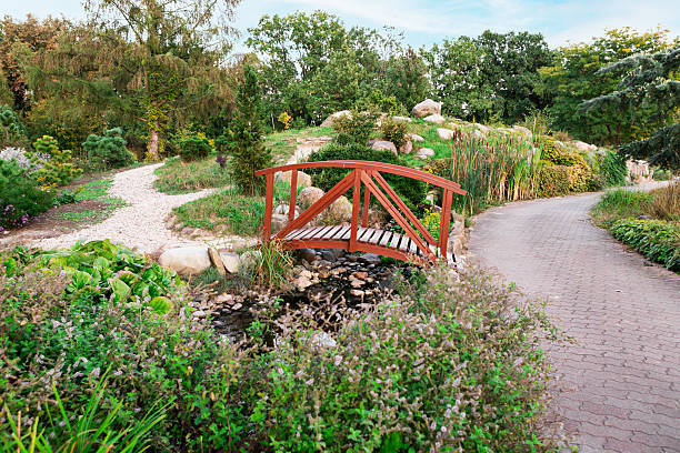 Wooden bridge over dry river with stones in park. Green lawn, bushes and trees in summer on sunny day. Background for design. Ecology and nature. Landscape design.
