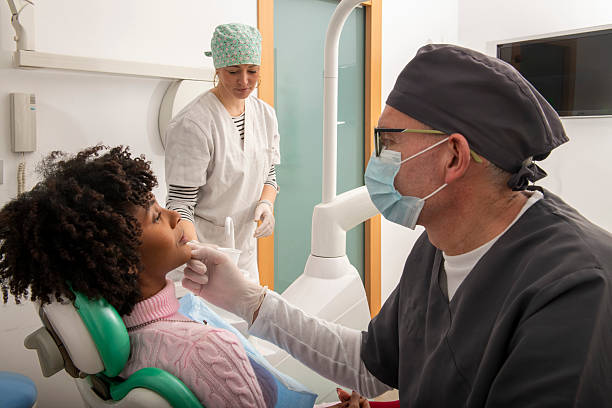 Dentist examining patient's teeth in dental clinic with assistant preparing tools
