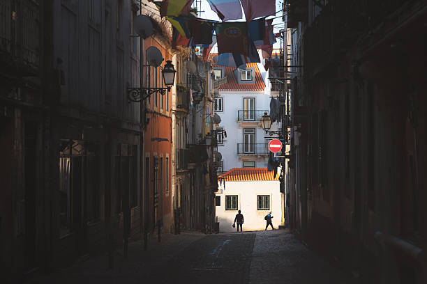 Lisbon, Portugal - July 15,2024: A moody, narrow alleyway in Bairro Alto, Lisbon, is cast in dramatic light and shadow, with silhouettes of people walking, capturing the gritty, historic atmosphere of the old town.