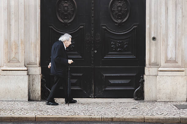 Lisbon, Portugal - July 15 2024: Quiet daily life scene in the old town as an elderly man with an umbrella as a cane strolls along a cobblestone sidewalk in Bairro Alto.