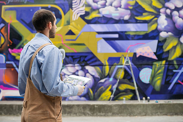 Rear shot of graffiti artist using his reference design to make a mural on a outdoor wall.