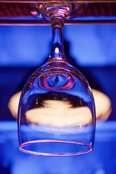 A close-up of an empty wine glass hanging upside down from a rack in a bar. The background is illuminated with blue and pink lights, creating a vibrant atmosphere.