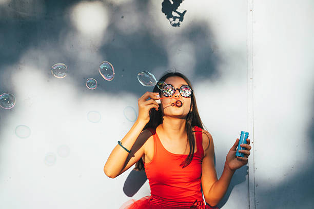 A radiant girl blows bubbles with a joyful smile, showcasing a blend of wonder and playfulness, dressed in a vibrant red outfit against a bright, sunny backdrop.