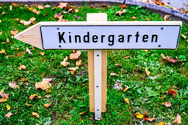 A wooden sign points towards a kindergarten in a park setting, with green grass and scattered yellow leaves. The sign adds a playful touch to the area, inviting young children and parents.