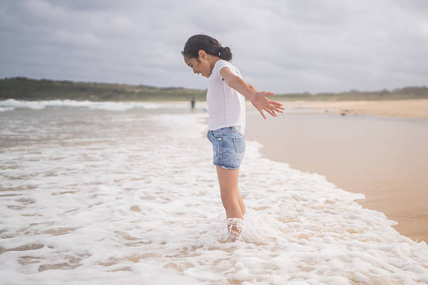 Pacific Islander little girl standing on the beach and letting waves cover her feet in shallow water.