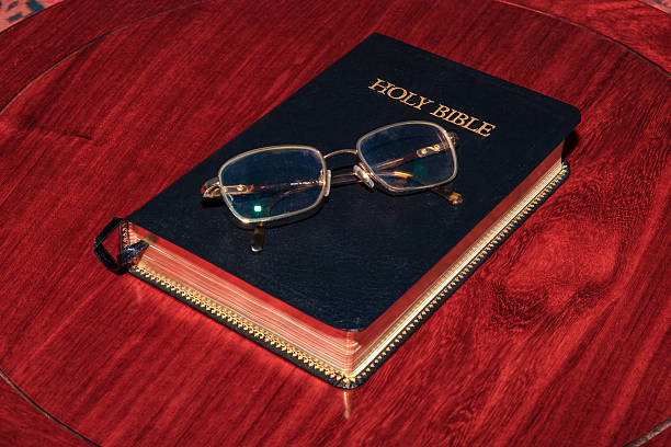 The Holy Bible placed on a richly grained rosewood table. The reader's eyeglasses have been placed on the cover, indicating the reader has taken a break. The Bible is closed. The bottom part of a ribbon bookmark is visible.  Indoor shot copy space. No people.