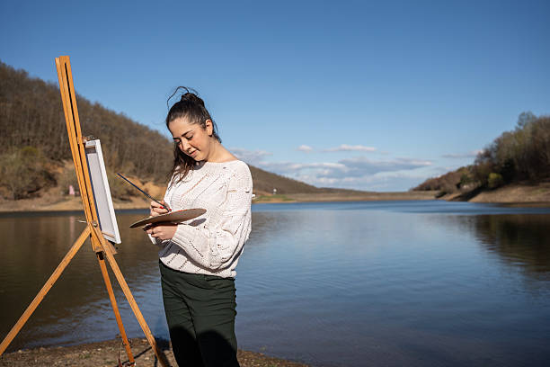 A creative individual enjoying outdoor painting by a calm lake, surrounded by nature, conveying peace and creativity. Bright sunlight and blue skies add to the serene and productive atmosphere.