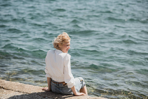 Against the backdrop of Opatija's sparkling sea, a blonde woman sits on the steps with her legs in the water, the wind playfully tousling her hair. The rhythmic waves and gentle breeze make for a truly tranquil scene.
