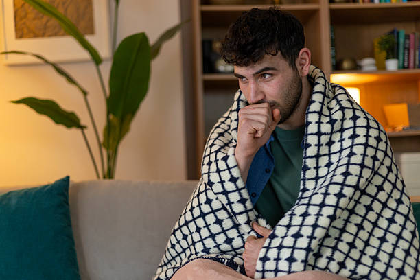 istock Sick young man coughing while sitting on the sofa at home, wrapped in a blanket 2209961202