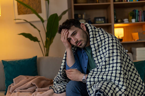 istock Sick young man suffering from a headache, wrapped in a blanket on the sofa, feeling unwell 2209961236