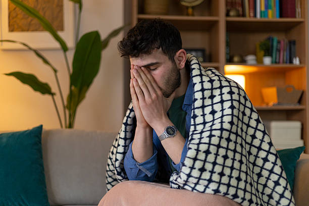 istock Sick young man suffering from cold and flu covering himself with a blanket while sitting on the sofa at home 2209961261